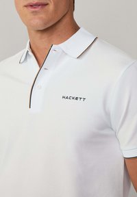 Polo bianco con colletto strutturato e piping blu contrastante, dotato di un piccolo logo ricamato "HACKETT" sul petto.
