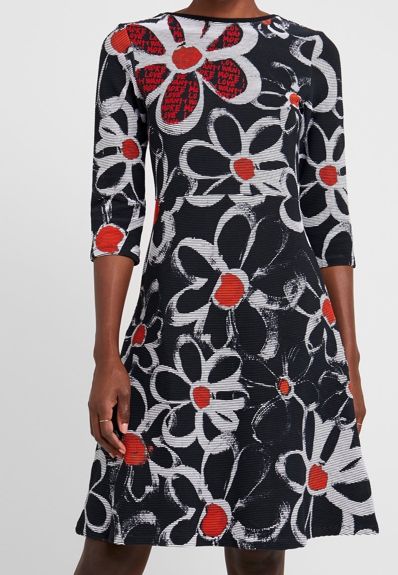 Robe noire avec de grandes fleurs blanches et des centres rouges, manches trois-quarts, portée par une personne montrée du cou aux genoux.