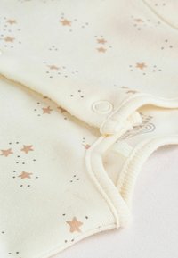 Combinaison jaune en tissu doux, ornée d'étoiles marron clair éparpillées et de petits points. Fermetures à pression au niveau du col et des bords des bras.