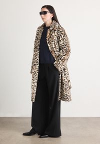 Kabát z imitácie leopardieho kožucha so šálovým golierom, nosený cez tmavý sveter a široké čierne nohavice, doplnený čiernymi slnečnými okuliarmi.
