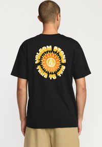 T-shirt noir à manches courtes avec un grand graphique jaune et rouge représentant un effet de rayon de soleil et le texte "VOLCOM STONE TRUE TO THIS" au dos.
