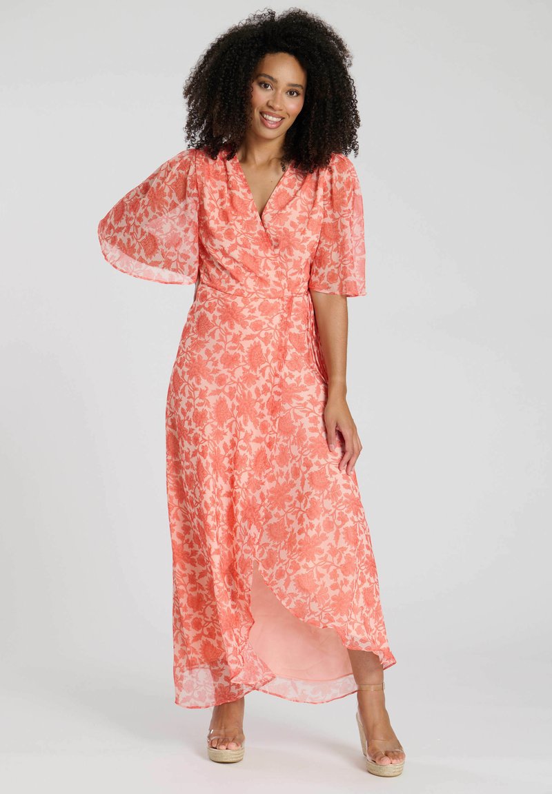 Maxikleid in Koralle mit floralem Muster, kurzen schwingenden Ärmeln, Wickeldesign und einem seitlichen Schlitz für zusätzlichen Schwung. Kombiniert mit beigen Plateausandalen.