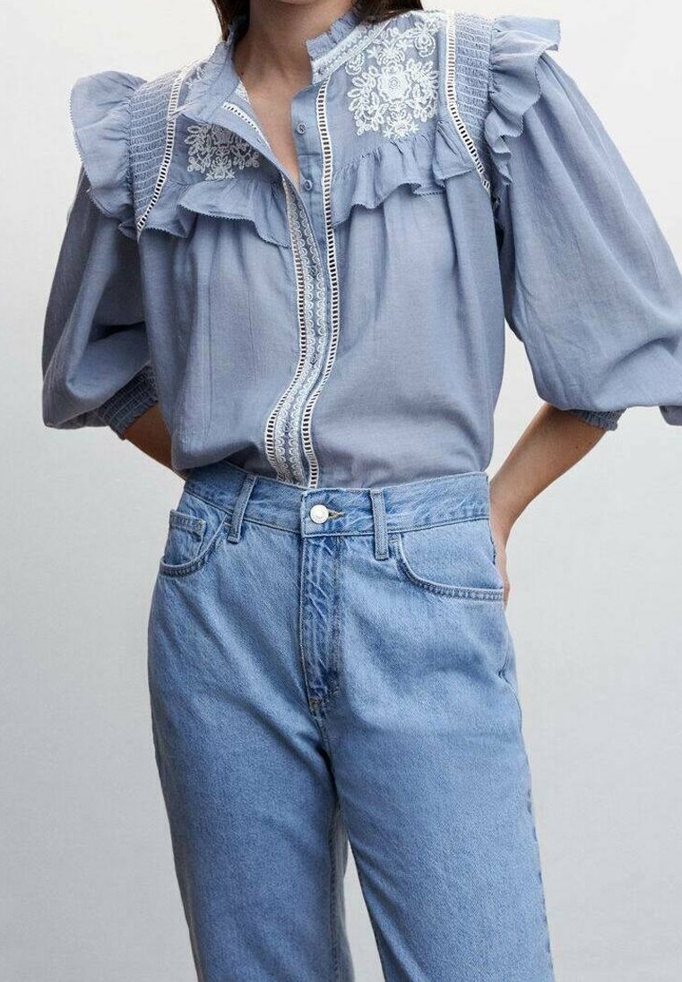 Femme portant un chemisier bleu clair avec des détails en dentelle et des volants, associé à un jean en denim bleu clair taille haute, sur un fond uni.