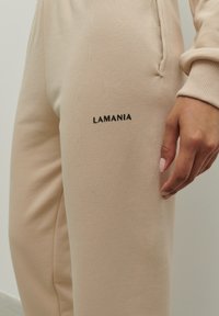 Béžové teplákové nohavice vyrobené z mäkkého materiálu, s čiernym logom "LAMANIA" na ľavom stehne a elastickým pásom a manžetami.