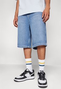 Lysblå denimshorts, knælange, med lige snit. Parret med sort-hvide sneakers og hvide sokker med gule og blå striber.