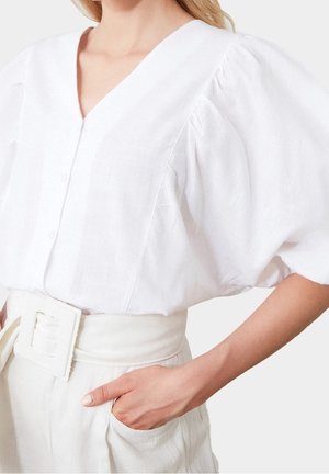 Frau trägt eine weiße Bluse mit V-Ausschnitt und Puffärmeln sowie eine weiße, hoch taillierte Hose mit Gürtel und einer Hand in der Tasche.