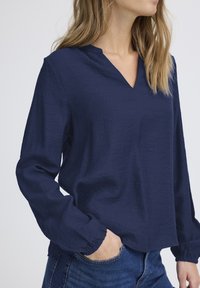 Blusa blu navy a maniche lunghe in tessuto strutturato con scollatura a V e polsini elasticizzati, abbinata a jeans in denim.
