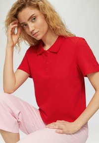Rode polo met korte mouwen met een klassieke kraag en knoopsluiting; model draagt lichtroze broek, gladde stof en aansluitend ontwerp.