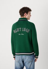 Groen gebreid sweater met een polokraag, voorzien van "GANT 1949" geborduurd op de achterkant. Ribgebreide manchetten met marineblauwe, witte en groene strepen.