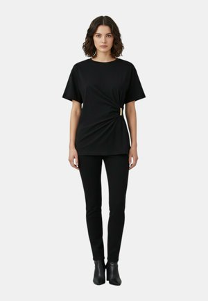 Mujer de pie con una blusa negra de manga corta con detalle fruncido a los lados y pantalones negros ajustados, combinados con botines negros.