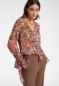 Blusa floreale marrone con scollatura a V, davanti arricciato e maniche a sbuffo. Presenta dettagli in rosa e crema con un motivo a stampa animale.