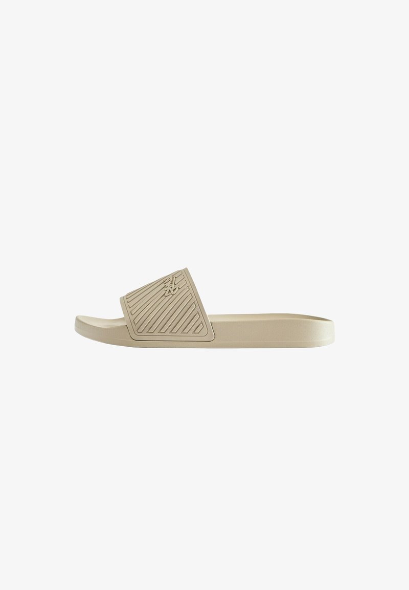 Sandale slide beige avec une large bride texturée présentant des lignes diagonales et un petit logo embossé sur fond blanc.