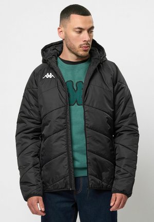 Zwarte pufferjack met een capuchon, ritssluiting en chevronstiksels. Kappa-logo in het wit op de linkerschouder. Draagt over een groene sweatshirt.