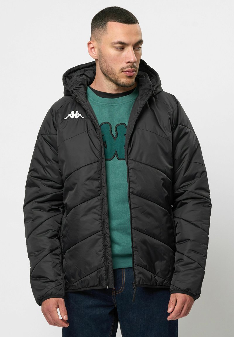 Schwarze Pufferjacke mit Kapuze, Reißverschluss und Chevron-Steppung. Kappa-Logo in Weiß auf der linken Schulter. Getragen über einem grünen Sweatshirt.