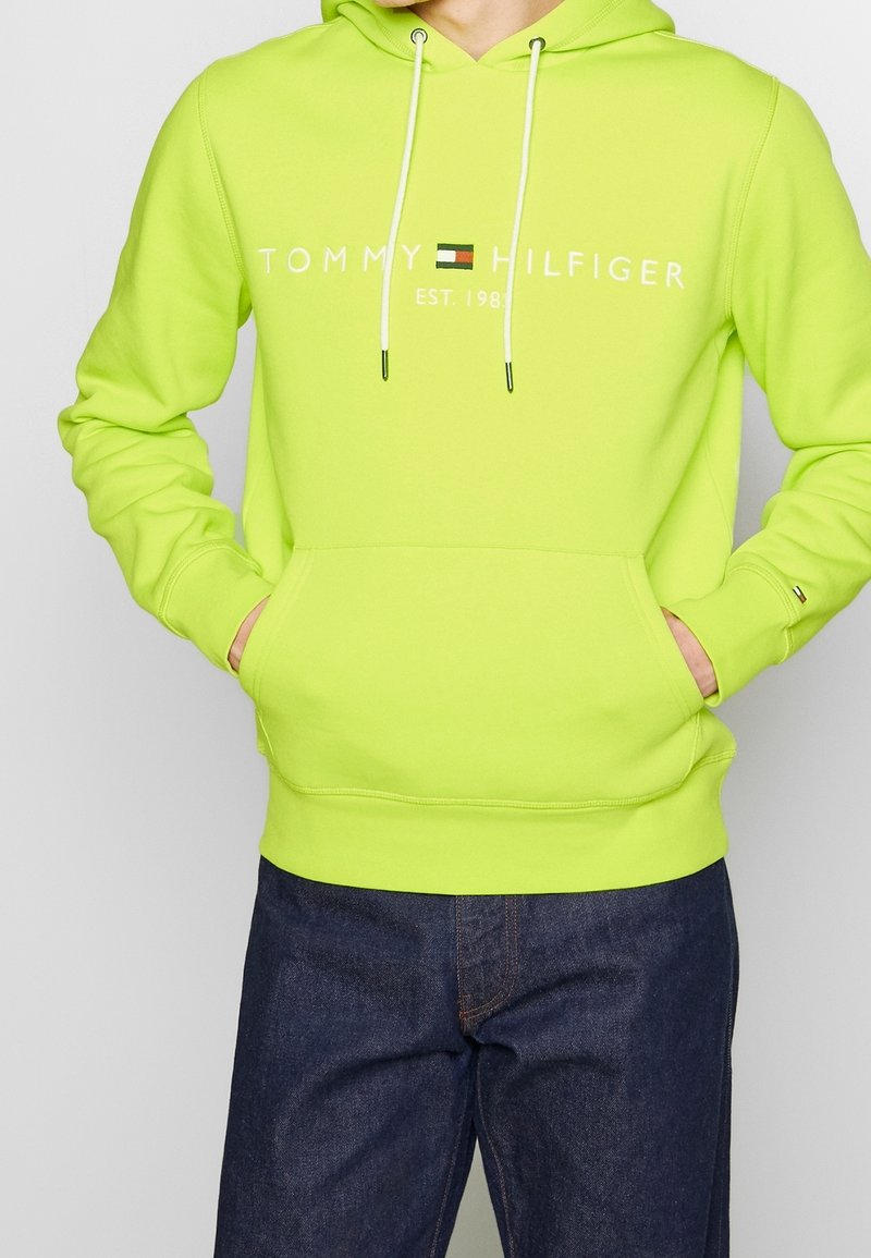Felpa verde neon con cordini, tasca frontale e logo bianco "TOMMY HILFIGER EST. 1985". Presenta polsini e orlo a coste.