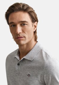 Pilkas polo marškinėlis iš tekstūruotos medžiagos, turintis vientisą kūną, trumpas rankoves, apykaklę ir mažą siuvinėtą logotipą ant krūtinės.