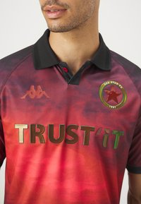 Polo shirt con gradiente rosso e colletto nero. Presenta il logo Kappa, il testo riflettente verde "TRUST'IT" e un emblema a stella rossa.