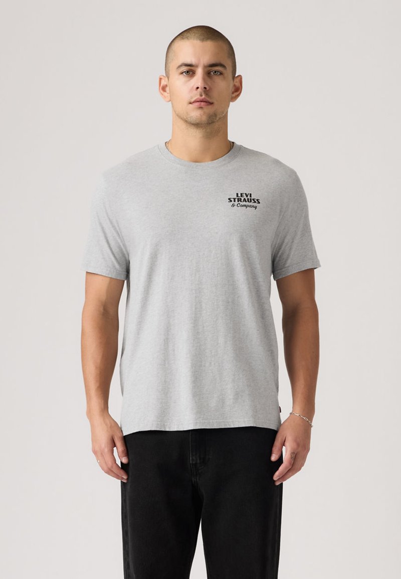 T-shirt en coton gris à manches courtes, col rond, avec un logo imprimé noir sur le haut à gauche de la poitrine. Coupe décontractée, texture lisse.