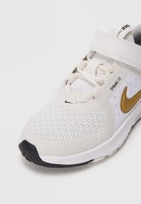Białe buty sportowe z siateczkowym przodem, wiązaniem i zapięciem na rzep, złotym logo Nike, napisem "Dynamic Fit" oraz czarnymi akcentami na podeszwie.