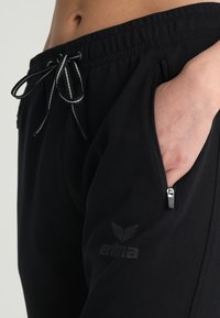 Pantaloni jogger neri realizzati in tessuto morbido, con vita regolabile con cordino, tasche con zip e un logo sulla coscia sinistra.