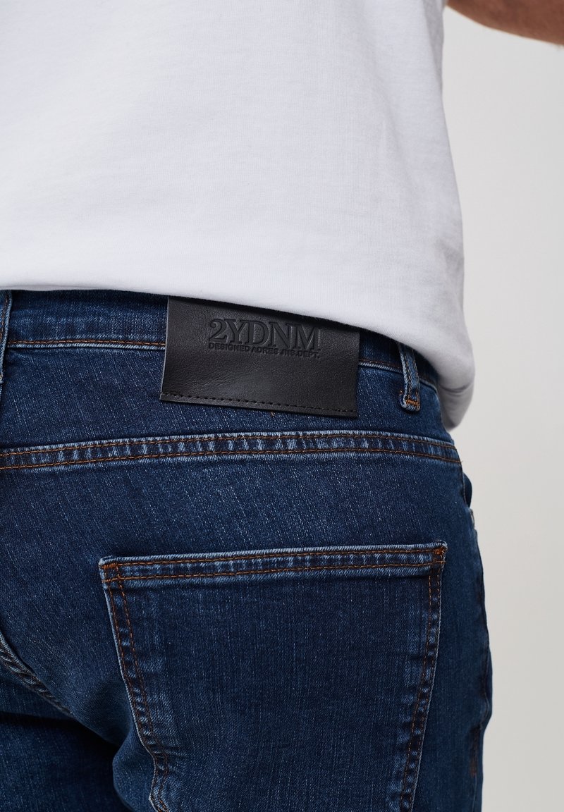 Vue de dos d'une personne portant un jean denim bleu foncé avec une étiquette en cuir noir sur la ceinture et une chemise blanche rentrée.