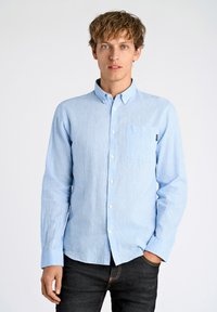 Camisa azul claro de manga larga y botones, hecha de tela texturizada, con un bolsillo en el pecho y un cuello clásico, combinada con jeans oscuros.