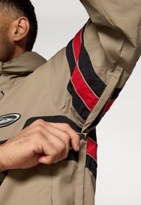 Καφέ σακάκι με μαύρες και κόκκινες ρίγες. Διαθέτει φερμουάρ και ρυθμιζόμενους καρπούς. Λείο ύφασμα με χαλαρή εφαρμογή. Ορατή ετικέτα λογότυπου.