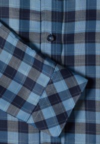 Tissu à carreaux bleu et noir avec des coutures visibles, montrant un gros plan du poignet d'une chemise et du bouton.
