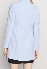 Donna che indossa un cappotto su misura di colore blu chiaro e una gonna nera, vista di spalle su uno sfondo semplice.