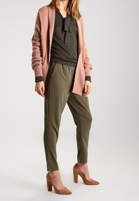 Ljusrosa cardigan, mörkgrön topp med knytdetalj, olivgröna byxor och beige högklackade skor. Mjuka texturer och figurnära design.