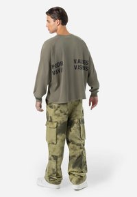 Sudadera verde oliva con mangas raglán que presenta texto negro en la parte posterior. Pantalones cargo con estampado de camuflaje, múltiples bolsillos y un ajuste relajado.