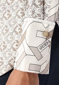 Long-sleeve shirt with a light fabric featuring a geometric beige and white pattern and a "GUESS" logo on the cuff. Gold button accent.