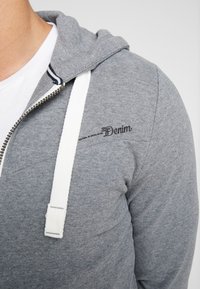 Sudadera gris con cremallera y cordones blancos. Presenta una textura de tejido ligero y un estampado negro de "Denim" en el pecho. Diseño clásico con capucha interior a rayas.