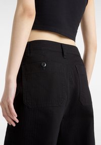 Svart högmidjad byxa, tillverkad av denim med en slät textur. Har två bakfickor med knapp, samt sömnadsdetaljer längs sömmen.