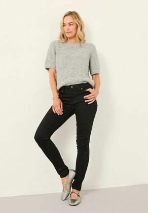 Kvinde iført en grå kortærmet sweater, sorte bukser og sølvfarvede flade sko, stående med det ene ben krydset og den ene hånd i lommen.