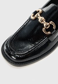 Mocasín negro de charol con un grueso adorno en cadena dorada y superficie texturizada. Punta redondeada y detalles de costura en contraste.