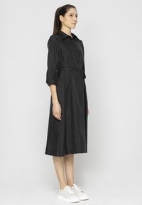 Czarna sukienka koszulowa z satyny, z kołnierzykiem, zwiniętymi rękawami o długości trzech czwartych i paskiem w talii, zestawiona z białymi sportowymi sneakersami na platformie.