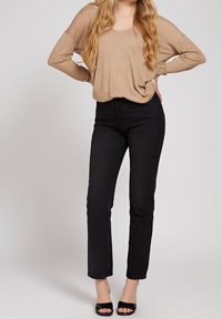 Top beige de manga larga con un corte holgado, combinado con unos jeans negros ajustados. Tacones negros de punta abierta completan el conjunto. Fondo sencillo.