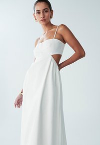 Vestido de lino blanco con un corsé ajustado, tirantes finos y detalle cortado en la cintura. Falda fluida que cae hasta el suelo.
