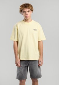 Yellow cotton t-shirt with short sleeves and a round neckline, featuring a black graphic print on the left chest, paired with gray denim shorts.