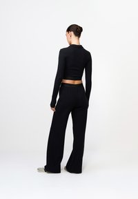 Mustade lühikeste varrukatega topp ja laiad püksid. Sile, venitav kangas, millel on nähtav vöökoht. Mudel on seljaga kaamera poole valge tausta ees.