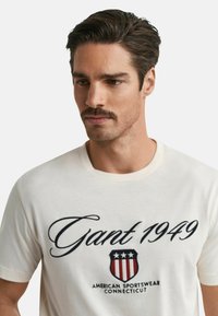 Cremefarvet T-shirt med en fed marineblå skrift "Gant 1949" og et skjoldlogo med røde og hvide striber, blødt bomuldsstof, rund halsudskæring.