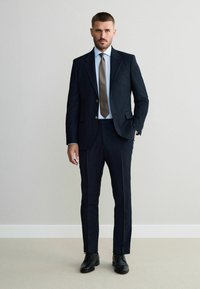 Navy pinstriped suit with a tailored jacket and matching trousers, paired with a light blue shirt and a textured tan tie. Black shoes.