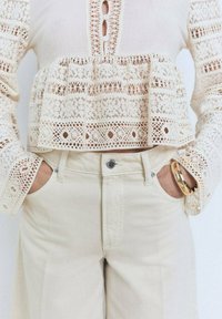 Top in pizzo beige chiaro con motivi intricati, tre bottoni sul collo, abbinato a pantaloni beige a vita alta e un bracciale dorato.