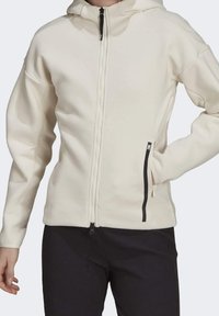 Světlobéžová mikina na zip s texturovaným povrchem, kapucí a černým zipem. Obsahuje boční kapsy s černými detaily. Hladký textilní povrch.