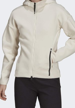 Haut de hoodie zippé beige clair avec une finition texturée, capuche et fermeture éclair noire. Comprend des poches latérales avec des accents noirs. Texture de tissu lisse.