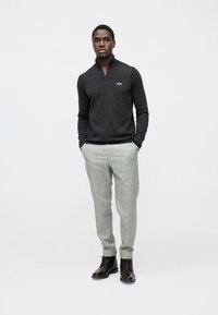 Sweatshirt preto com fecho zip e logo branco, combinado com calças cinza claro às riscas finas e botas pretas com atacadores, em pé contra um fundo neutro.