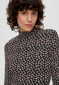 Kobieta z kręconymi włosami, ubrana w czarną bluzkę z wysokim kołnierzem w mały beżowo-czerwony wzór w panterkę na jednolitym tle.