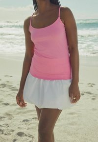 Débardeur rose en tissu léger associé à une jupe blanche courte et évasée. Plage de sable et océan en arrière-plan.