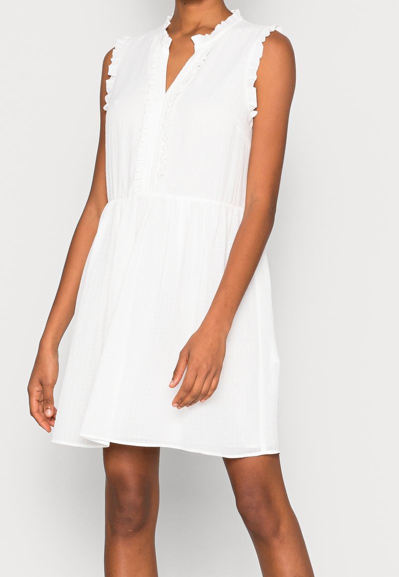 Robe blanche sans manches avec des bords volantés aux épaules et un décolleté en V, portée par une personne debout les bras détendus le long du corps.
